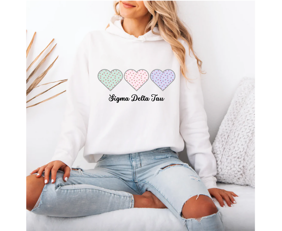 White hoodie with colorful heart designs and 'Sigma Delta Tau' text, worn by a person sitting on a white couch.