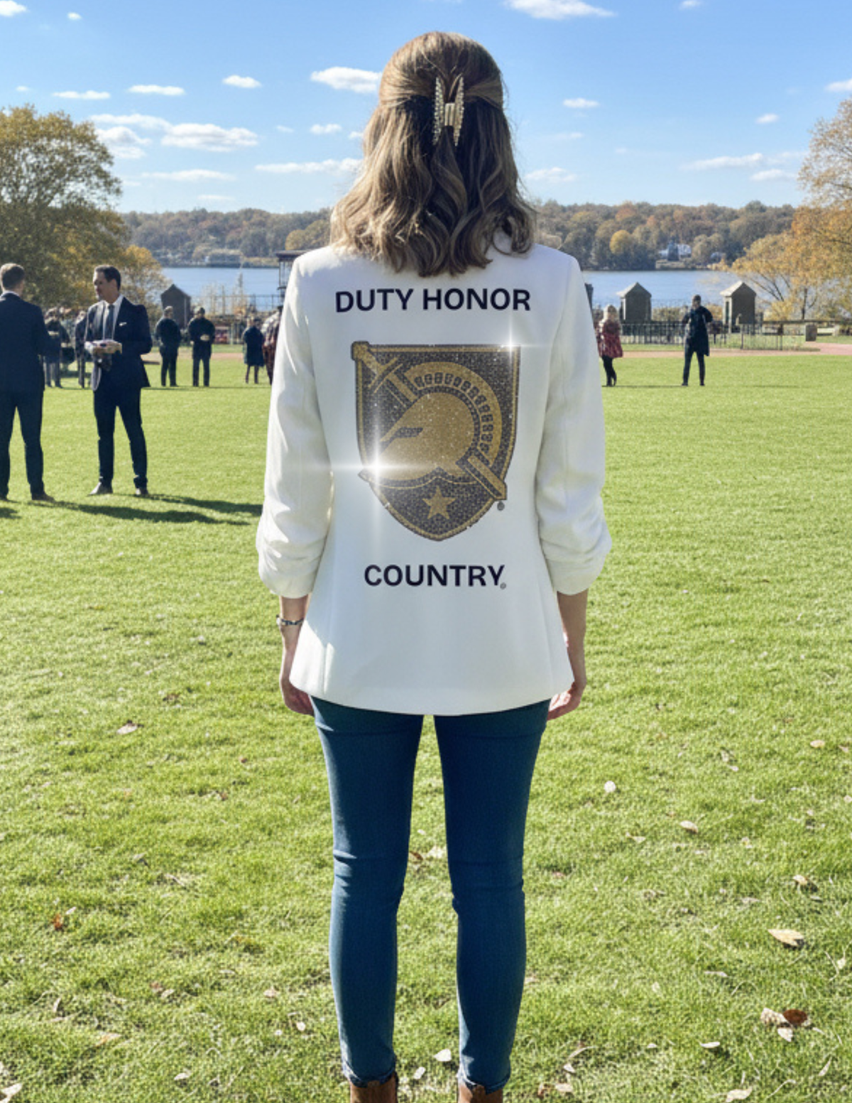 A woman standing outdoors on a grassy park lawn, photographed from behind, wearing a white long-sleeve top with 'DUTY HONOR' and a large gold-toned crest graphic on the back, paired with dark blue jeans.