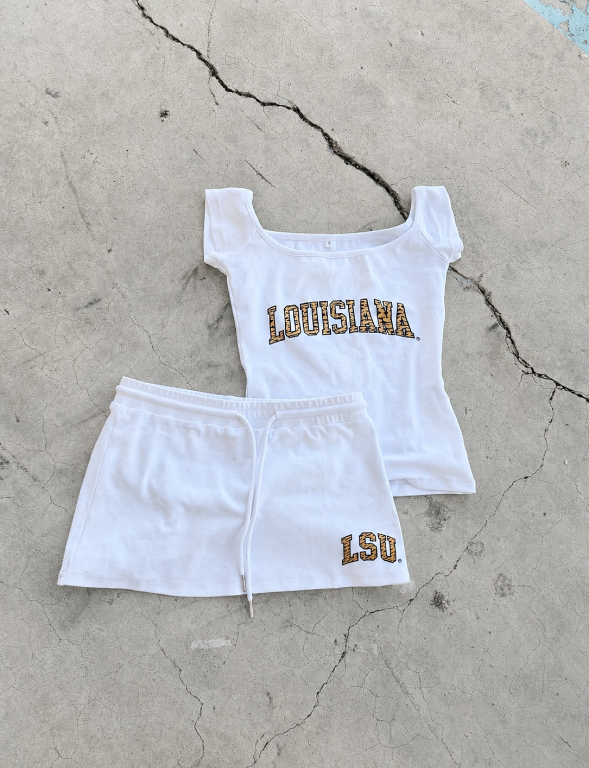 A white two-piece athletic set consisting of a scoop-neck tank top and matching shorts, laid flat on a cracked concrete surface. The top features 'LOUISIANA' in gold glitter lettering, and the shorts feature 'LSU' in a similar gold glitter font.