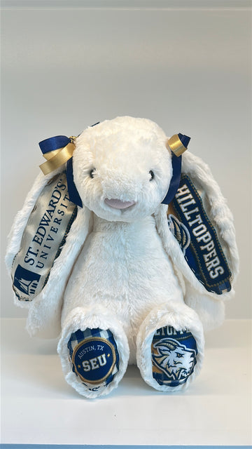 A white plush bunny toy with long floppy ears, decorated with St. Edwards University themed ribbons on the ears and blue-and-white patterned fabric on the feet, photographed against a plain light background.
