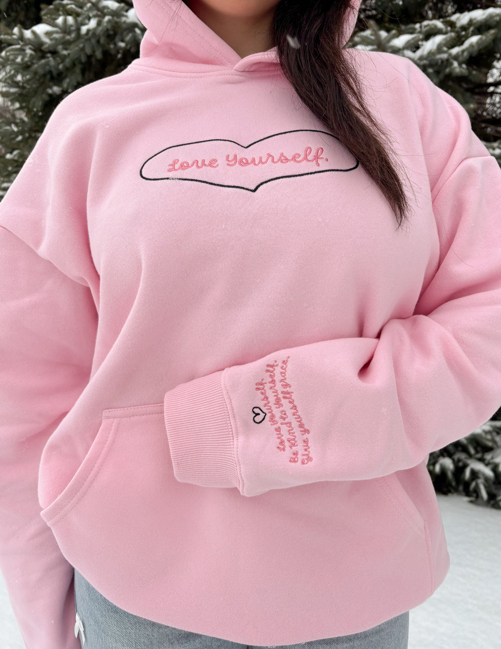 Pink hoodie with 'love yourself' text worn by a person outdoors.