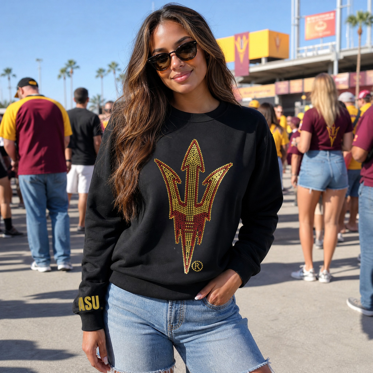 A woman wearing a black crewneck sweatshirt with a large Arizona State Sun Devils logo on the front, paired with light-wash denim shorts, standing outdoors at what appears to be a stadium or event venue.