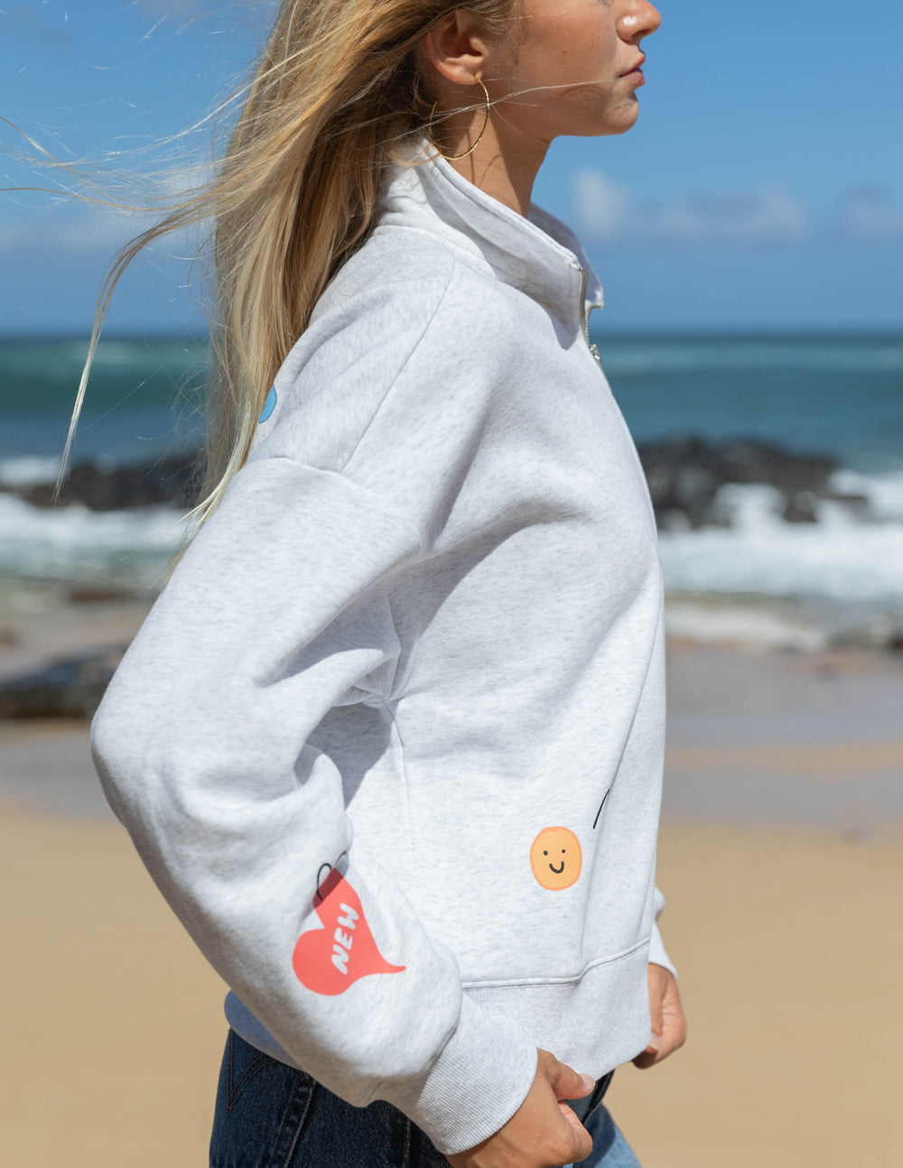 Person wearing a light gray hoodie with colorful patches on a beach