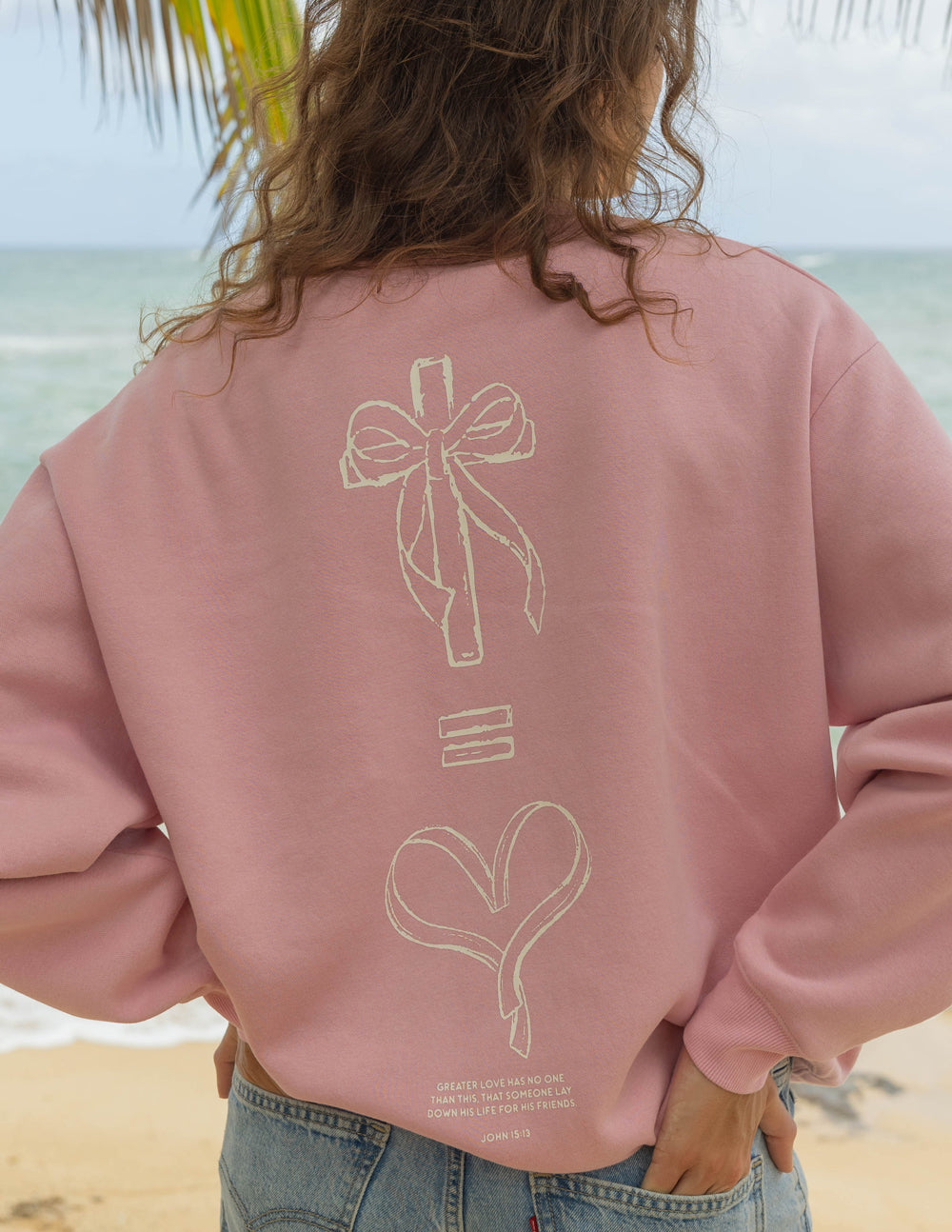 Person wearing a pink sweatshirt with a design on the back, standing on a beach.