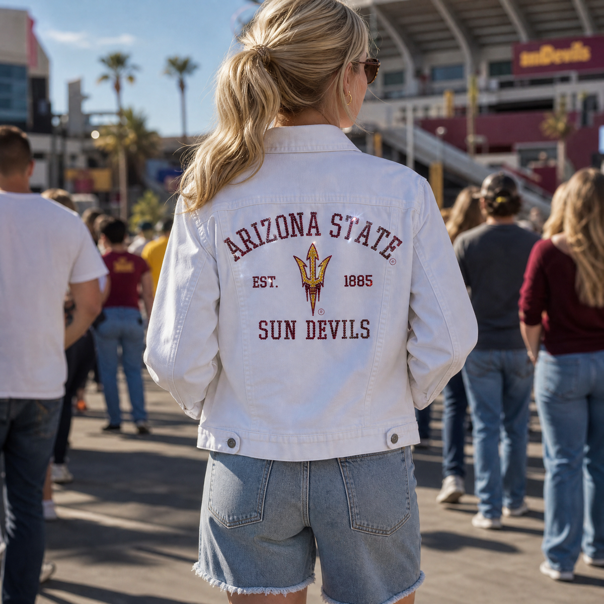 A woman viewed from behind wearing a white denim jacket with 'ARIZONA STATE SUN DEVILS EST. 1865' and the team logo printed on the back, standing in a crowded outdoor stadium setting.