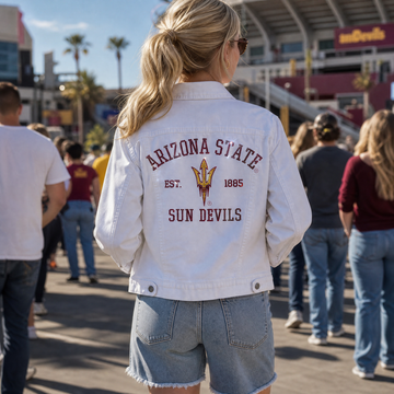 A woman viewed from behind wearing a white denim jacket with 'ARIZONA STATE SUN DEVILS EST. 1865' and the team logo printed on the back, standing in a crowded outdoor stadium setting.