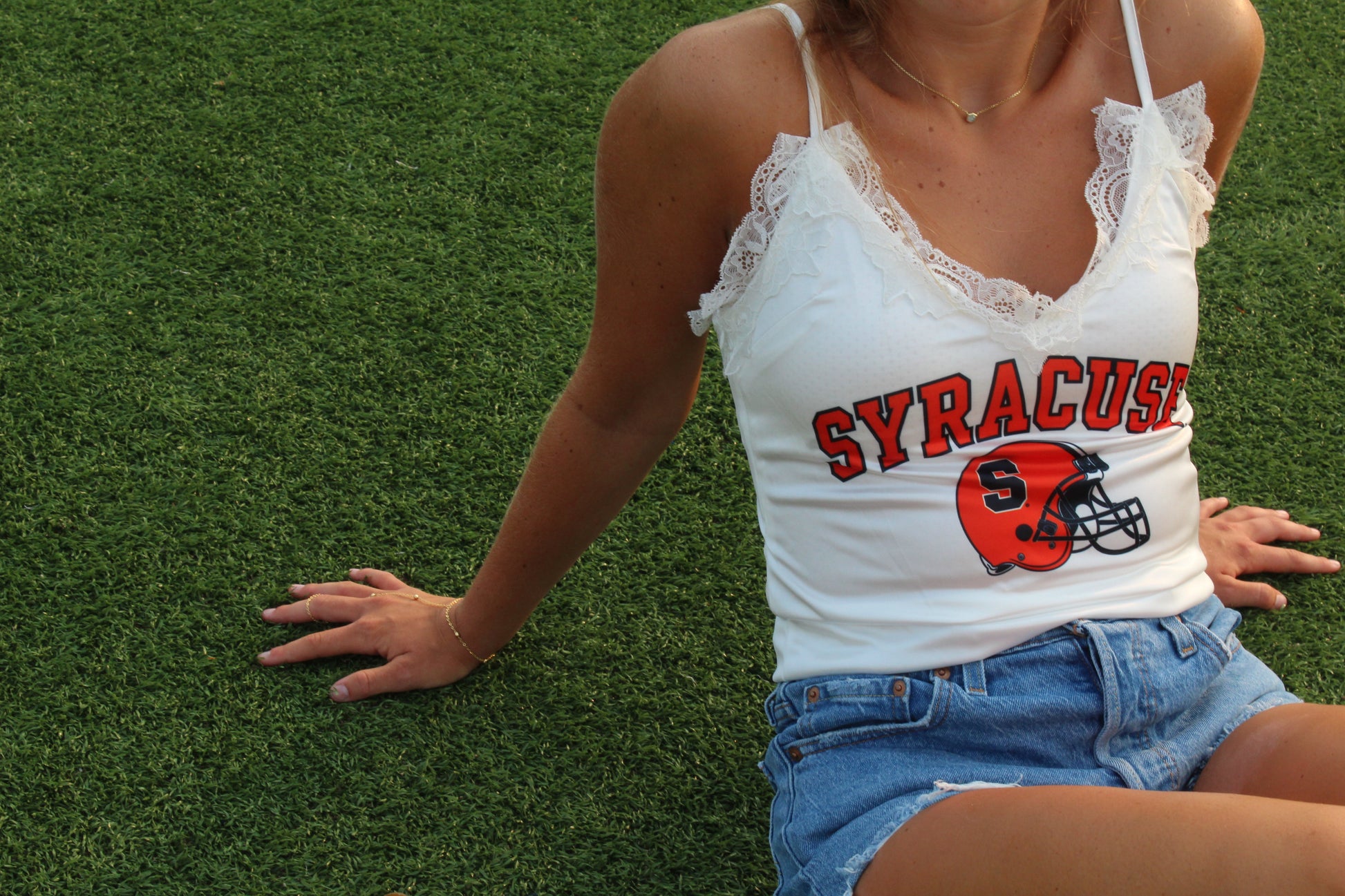 A woman sitting on green grass wearing a white lace-trimmed spaghetti-strap tank top with 'SYRACUSE' and a football helmet graphic in red, orange, and navy, paired with denim shorts.