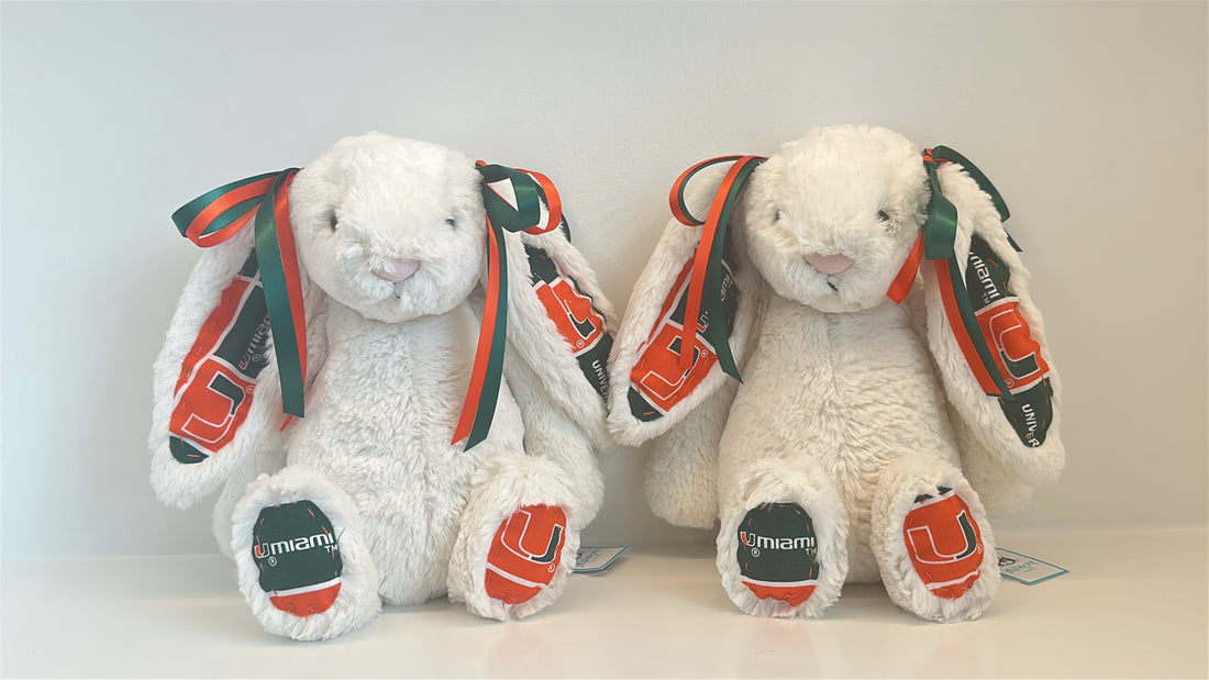 Two Cream Jellycats that are adorned with orange and white bows, University of Miami print on their ears and paws.