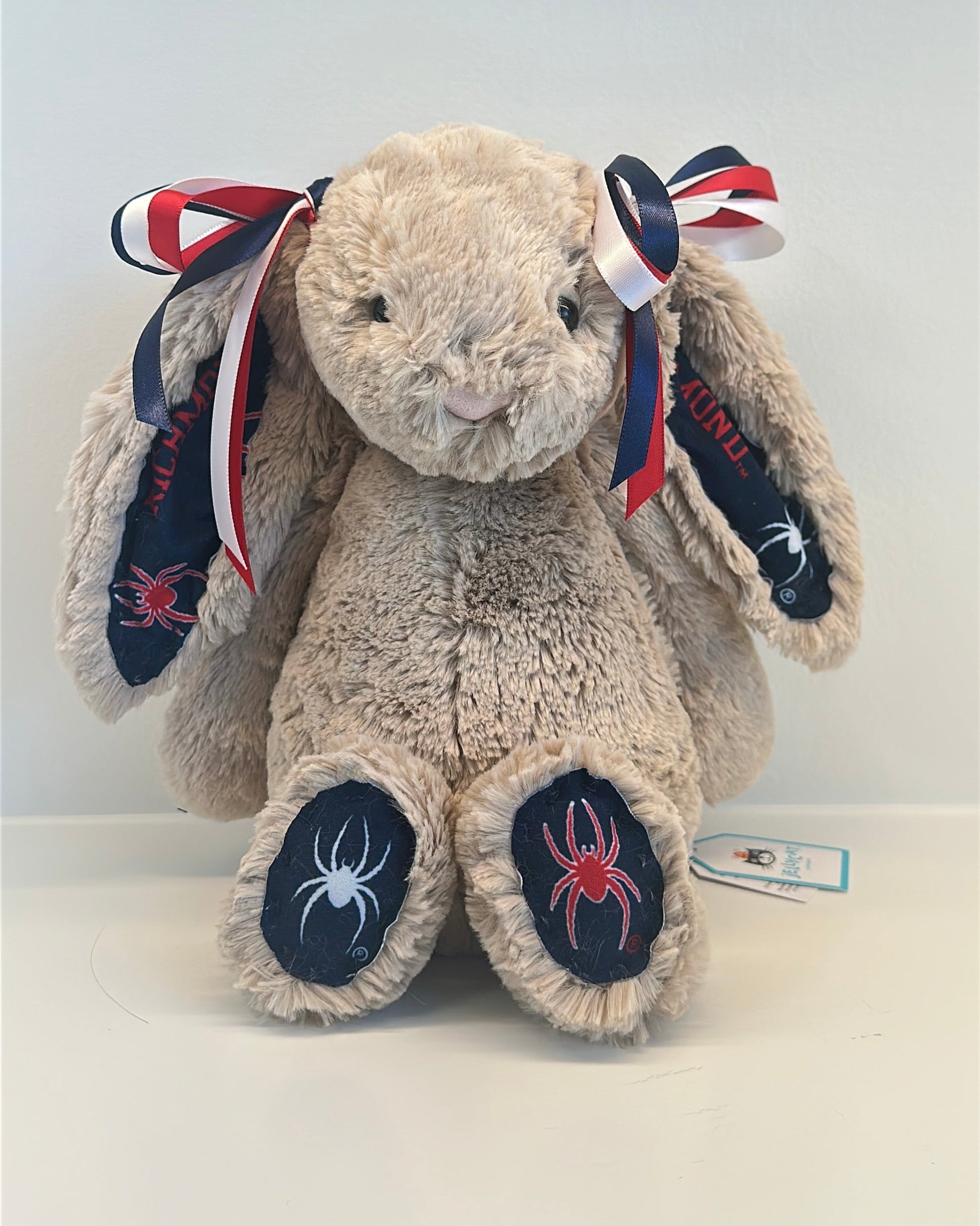 Plush rabbit toy with spider design on feet and red, white, and blue ribbons on a white background