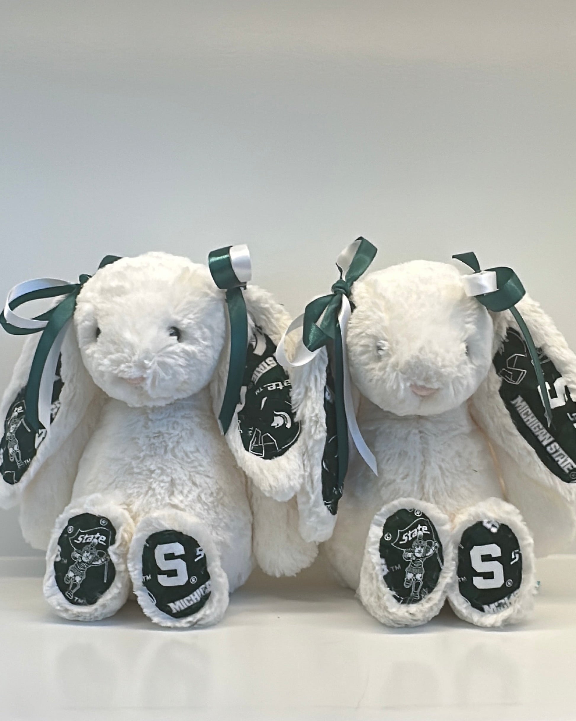 Two white stuffed animals with black ribbons and 'S' logos on a white background michigan state