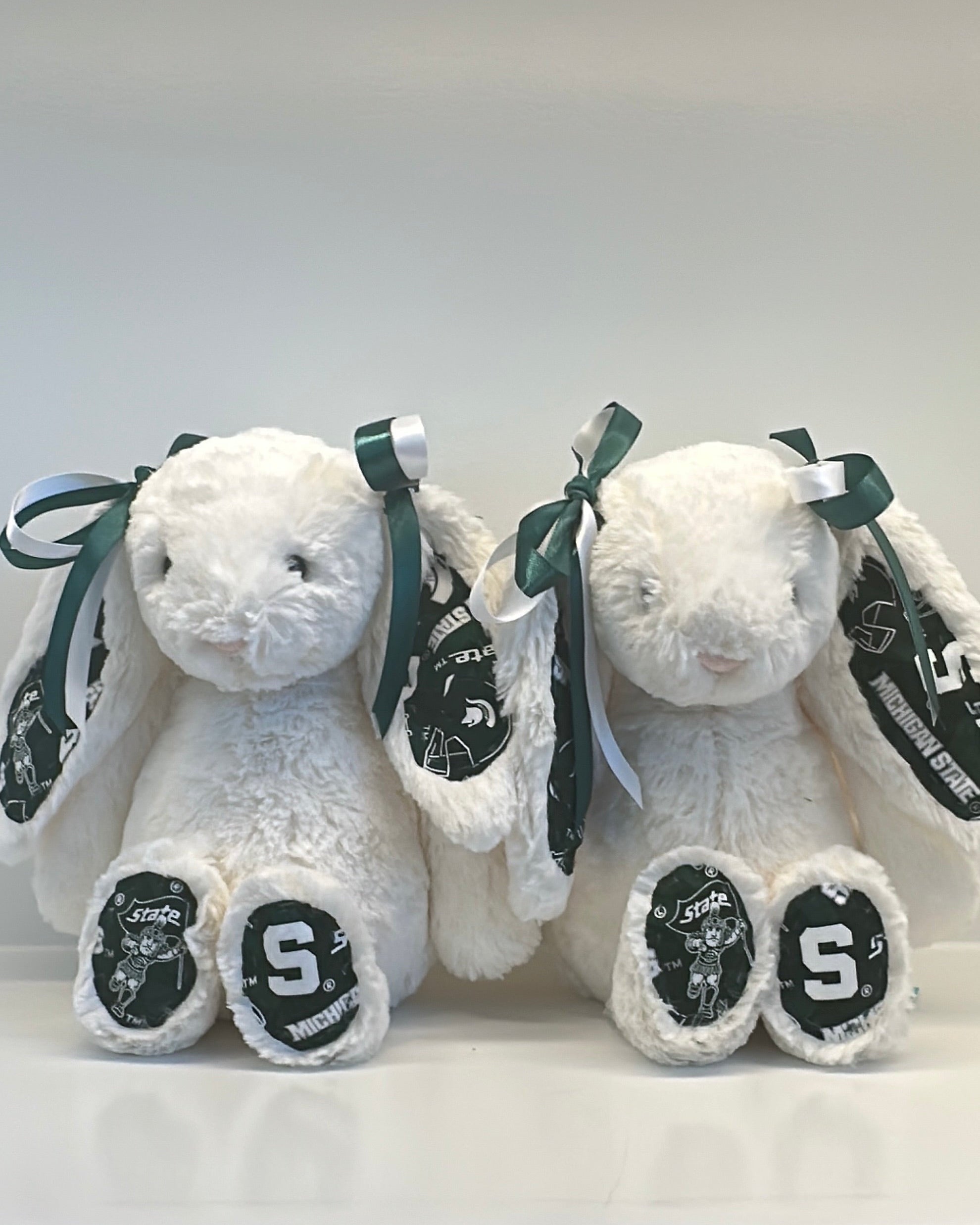 Two white stuffed animals with black ribbons and 'S' logos on a white background michigan state
